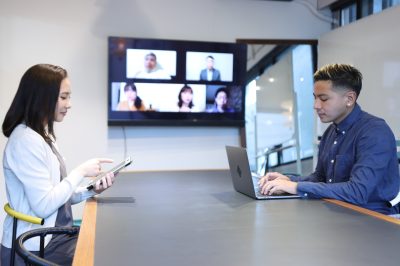 Employees in an office boardroom are joined by participants joining by video call